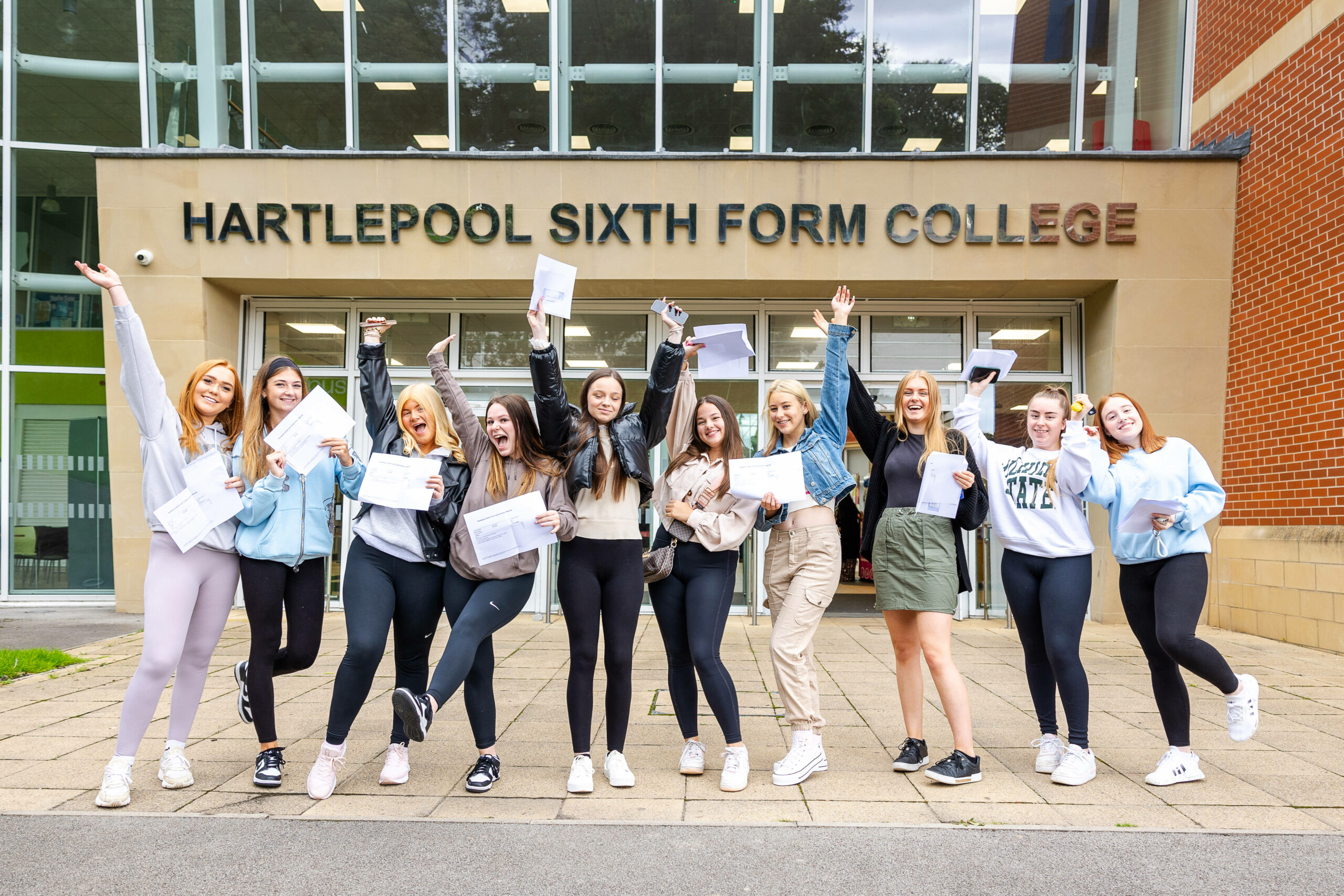 A group of students, holding their GCSE results, smile in front of the Hartleepool Sixth Form building.